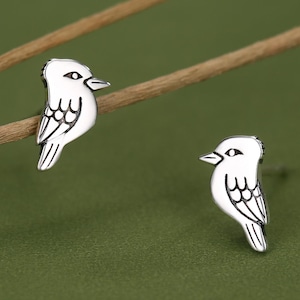 May include: A pair of silver stud earrings shaped like small birds. Each earring features a polished silver finish with engraved black lines detailing the bird's eye, beak, and wing feathers. The earrings are designed as post-back studs.