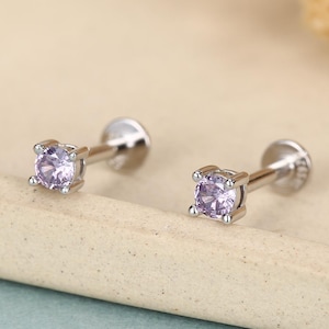 Può includere: Un paio di orecchini a lobo in argento con pietre preziose quadrate, viola chiaro. Gli orecchini sono presentati su una superficie azzurra, con fiori secchi sullo sfondo. Gli orecchini hanno un design a vite.