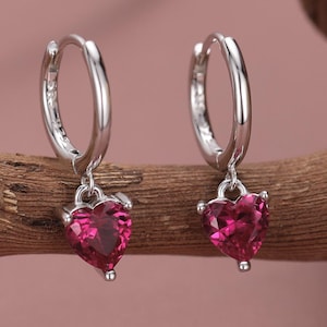 Op de afbeelding: Een paar zilveren oorringen met hartvormige, robijnrode hangers. De oorbellen worden gepresenteerd op een stuk bruin hout tegen een zachtroze achtergrond. De hartvormige hangers geven de sieraden een romantische uitstraling.