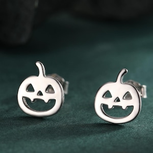 May include: A pair of silver pumpkin stud earrings. Each earring has a carved jack-o'-lantern face and a stem. The earrings are presented on a dark green surface, ideal for Halloween.