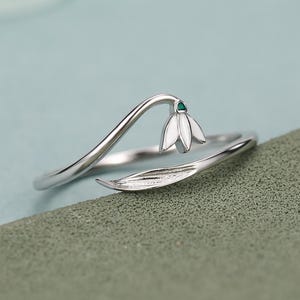May include: A silver-toned ring featuring a delicate, curved stem design that culminates in a white enamel flower bud with a tiny green accent at the centre. The band extends into a slender, pointed leaf shape that wraps around the finger.