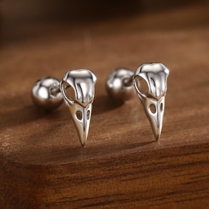 Op de afbeelding: Een paar zilverkleurige oorstekers in de vorm van vogel schedels. De oorbellen worden gepresenteerd op een donkerbruin houten oppervlak. De oorbellen hebben een gepolijste afwerking en zijn ontworpen met ingewikkelde details.