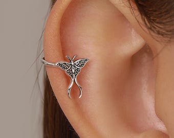 Può includere: Un ear cuff color argento con un design dettagliato a forma di falena con ali allungate e fluide. Il metallo presenta una finitura ossidata che mette in risalto i motivi intricati sulle ali, posizionato sul bordo esterno della cartilagine.