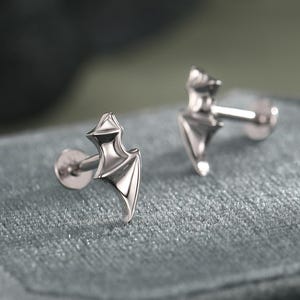 May include: A pair of silver-colored bat-shaped stud earrings. The earrings are displayed on a gray velvet surface. The bat design features pointed wings and a detailed body. The earrings have a post and backing for secure wear.