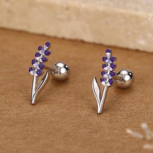 May include: A pair of silver-toned stud earrings featuring a lavender flower design with purple and white enamel detailing. The earrings have a post and ball closure, set against a neutral background.