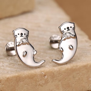 May include: A pair of silver-toned stud earrings shaped like small otters. Each earring features a polished metallic finish with engraved black details for the eyes, nose, and whiskers, depicting an otter holding a small round object in its paws.