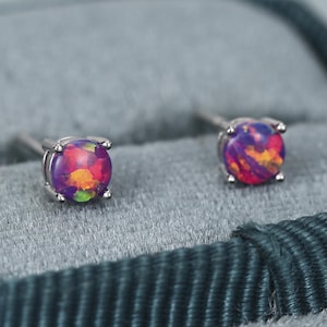 Peut inclure: Une paire de boucles d'oreilles à clous ronds avec des pierres d'opale synthétique aux reflets violets et orange, serties dans des griffes en métal argenté. Les boucles d'oreilles reposent sur une surface en velours gris avec une bordure en tissu.