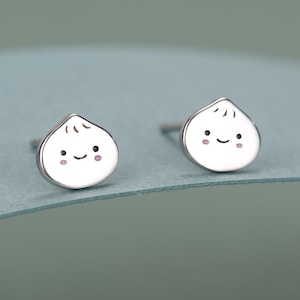 Könnte beinhalten: Ein Paar silberne Ohrstecker, jeweils geformt wie kleine, weiße Knödel mit einem lächelnden Gesicht. Die Knödel haben eine kleine, abgerundete Spitze und eine leicht strukturierte Oberfläche. Die Ohrringe sind vor blauem Hintergrund platziert.