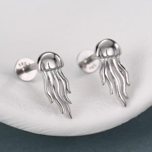 May include: A pair of silver-toned stud earrings shaped like jellyfish, each featuring a rounded top and multiple dangling tentacles. The earrings have a flat back post and are stamped with the text 585 on the base of the post.