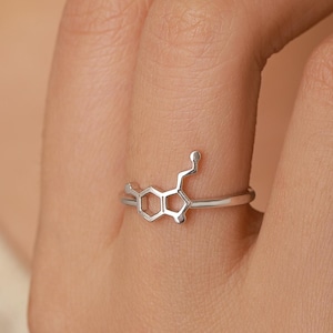 May include: A silver-coloured ring featuring a delicate, open-work chemical structure design on the front. The band is thin and polished, showcasing a molecular motif that sits prominently across the finger, crafted with a smooth, metallic finish.
