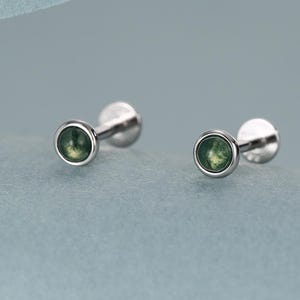 May include: A pair of small, silver-colored stud earrings with a round, green gemstone set in each. The earrings are displayed on a light blue surface. The earrings have a flat back and a small, round gemstone.