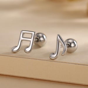 Peut inclure: Une paire de boucles d'oreilles argentées en forme de notes de musique. L'une est une noire, l'autre une croche. Les boucles d'oreilles ont une finition polie et un petit dos rond. Le fond est beige neutre.