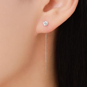 Puede incluir: Primer plano de una oreja con un pendiente de botón brillante y un pendiente de hilo de cadena plateada. El pendiente de botón es una piedra pequeña, redonda y transparente. El pendiente de cadena cuelga del lóbulo.