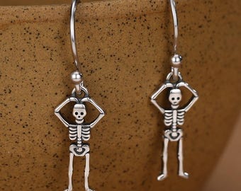 Può includere: Un paio di orecchini pendenti color argento con un ciondolo a forma di scheletro dettagliato. Ogni figura di scheletro ha le braccia sollevate sopra la testa, formando una forma circolare. Gli orecchini sono fissati a semplici ganci in metallo.