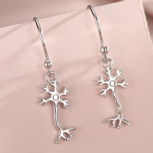 May include: Silver-coloured neuron-shaped drop earrings. The earrings depict a detailed neuron design, with a central cell body and branching dendrites and axon terminals. The earrings are displayed on a pink surface.