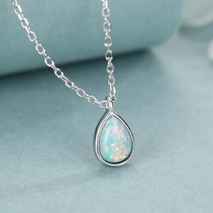 Puede incluir: Un collar de plata con un colgante en forma de lágrima que contiene una piedra de ópalo azul claro. El ópalo muestra destellos iridiscentes de color rosa y naranja en su superficie. El colgante cuelga de una delicada cadena de plata.