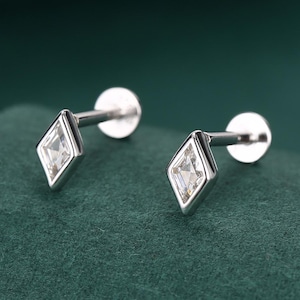 Può includere: Un paio di orecchini a lobo in argento. Ogni orecchino presenta una gemma trasparente a taglio quadrato incastonata in una cornice a forma di diamante. Gli orecchini sono esposti su una superficie verde scuro.