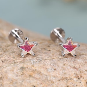 Puede incluir: Un par de pendientes de botón en tono plateado con un diseño de estrella de cinco puntas. Cada estrella tiene una piedra preciosa rosa iridiscente y facetada en el centro. Los pendientes cuentan con un poste trasero plano y circular.
