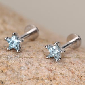 May include: A pair of silver-toned labret stud earrings featuring a five-pointed star-shaped setting. Each earring is set with a light blue, faceted gemstone at the centre of the star, attached to a straight metal post with a flat circular back.