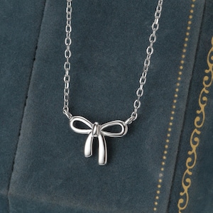May include: A silver necklace with a bow pendant. The bow is the focal point, suspended from a fine chain. The necklace is presented against a dark teal velvet jewellery box, which enhances the silver's gleam. A simple, elegant design.