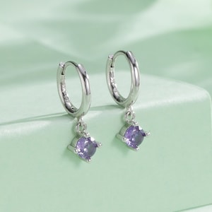 May include: A pair of silver hoop earrings with a small purple gemstone dangling from each hoop.
