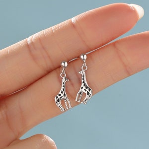 Könnte beinhalten: Ein Paar silberne Ohrhänger mit Giraffenmotiv. Die Giraffen sind mit schwarzen Punkten verziert und haben oben eine silberne Kugel.