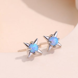 May include: A pair of silver stud earrings. Each earring has a starburst design with pointed edges and a light blue opal centre. The earrings are displayed on a light peach surface.