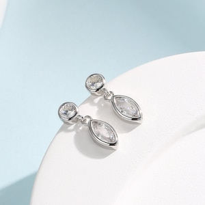 Puede incluir: Un par de pendientes de plata con piedras transparentes en forma de marquesa. Los pendientes tienen una pequeña piedra redonda en la parte superior y una piedra más grande en forma de marquesa que cuelga debajo.