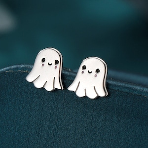 Könnte beinhalten: Ein Paar silberne Ohrringe in Geisterform mit lächelnden Gesichtern. Die Ohrringe befinden sich auf einer dunkelblauen Oberfläche.