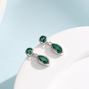 Puede incluir: Un par de pendientes de plata con gotas de piedras preciosas verdes. Los pendientes tienen una pequeña piedra preciosa redonda en la parte superior y una piedra preciosa más grande de forma de marquesa que cuelga debajo.