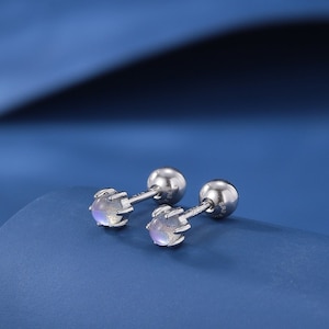 May include: A pair of silver stud earrings with a small, iridescent moonstone set in each. The earrings are on a blue surface.