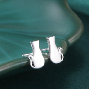 May include: A pair of silver cat-shaped stud earrings. The earrings are displayed on a teal-colored, leaf-shaped dish. The earrings have a polished finish and a simple, elegant design. The earrings are small and delicate, perfect for everyday wear.