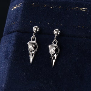 Puede incluir: Un par de pendientes de plata con un pequeño diseño de cráneo de pájaro que cuelga de cada botón. Los pendientes se muestran sobre una superficie de terciopelo azul oscuro.