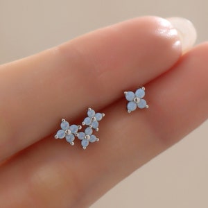 May include: A pair of silver flower stud earrings with light blue enamel. The earrings are shown on a person's hand.