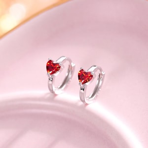 Puede incluir: Un par de pendientes de aro de plata con una piedra preciosa roja en forma de corazón en cada aro.