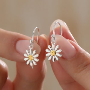 May include: Silver dangle earrings with a small daisy design. The center of the daisy is gold and has a smiley face.