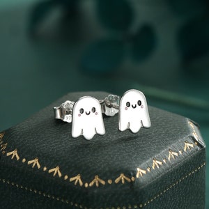 Può includere: Un paio di orecchini a lobo in argento con un piccolo design a forma di fantasma bianco con un viso sorridente. Gli orecchini sono esposti in una scatola di velluto verde.