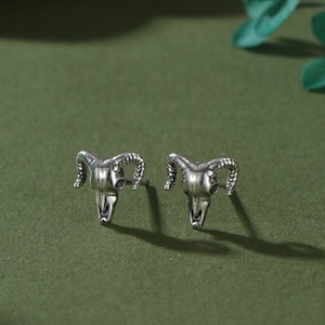 May include: A pair of silver earrings featuring a ram skull design. The earrings are displayed on a green surface.