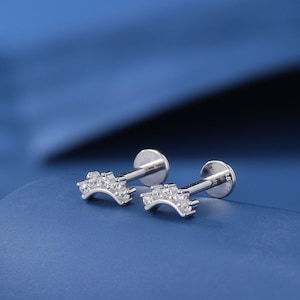 Può includere: Due orecchini a barra curva in argento con piccole pietre preziose trasparenti incastonate nel metallo. Gli orecchini sono su una superficie blu.