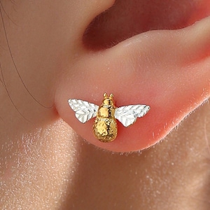 Puede incluir: Un par de pendientes de botón con forma de abeja, dorados y plateados. Los pendientes presentan un cuerpo dorado texturizado y alas plateadas, con un diseño detallado de insecto. Pequeños y delicados, son adecuados para el uso diario.