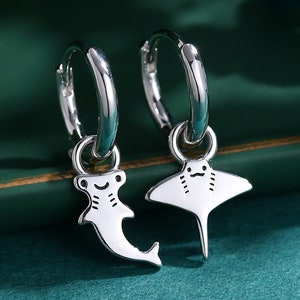 Puede incluir: Un par de pendientes de aro de plata con un tiburón martillo y una manta raya colgando de cada aro.