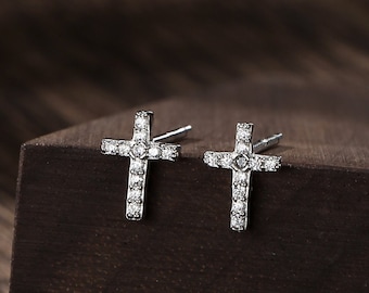 Puede incluir: Un par de pendientes de cruz plateados con pequeñas piedras de circonita cúbica transparentes engastadas en el diseño de la cruz. Los pendientes se muestran sobre una superficie de madera marrón oscura.