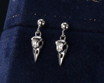 Puede incluir: Un par de pendientes de plata con un pequeño diseño de cráneo de pájaro que cuelga de cada botón. Los pendientes se muestran sobre una superficie de terciopelo azul oscuro.