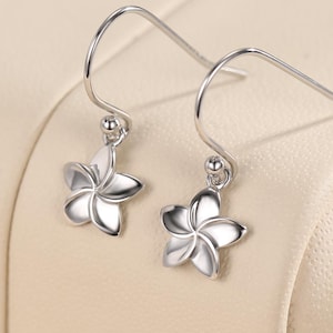 Puede incluir: Un par de pendientes plateados con forma de flor. Cada pendiente presenta un colgante de flor de cinco pétalos suspendido de un gancho. Los pendientes se exhiben sobre una superficie beige claro, mostrando su delicado diseño y brillo.
