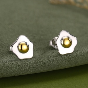 May include: A pair of silver stud earrings in the shape of fried eggs. Each earring has a white enamel egg white and a gold-coloured yolk. The earrings are presented on a green surface.