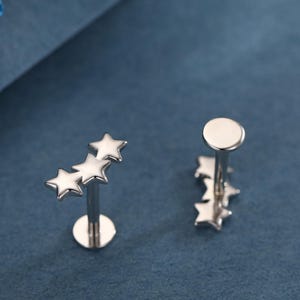 May include: Two silver-colored star-shaped earrings on a blue background. One earring features three stars, and the other has a single star design. The earrings have a flat disc backing.