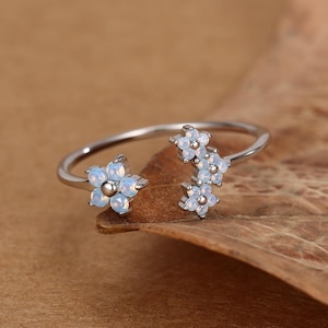Könnte beinhalten: Ein silberfarbener Ring mit floralem Design. Der Ring hat ein zartes Band, das sich teilt, um drei kleine, hellblaue Blumencluster zu präsentieren. Die Blumen haben einen subtilen, perlmuttartigen Glanz. Der Ring ist vor einem braunen Hintergrund platziert.