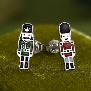 Può includere: Un paio di orecchini a lobo in argento con un colorato design a soldatino schiaccianoci. Un orecchino è verde e nero, l'altro è rosso e nero. Gli orecchini sono appoggiati su una superficie di velluto verde.