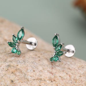 May include: Two silver-toned leaf-shaped earrings with vibrant green gemstones. Each earring features a cluster of marquise-cut and round-cut green stones, arranged to resemble leaves. The earrings have a post and backing for secure wear.