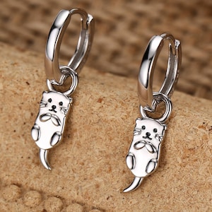Peut inclure: Boucles d'oreilles créoles argentées avec des breloques de loutre blanches et noires. Les loutres ont des visages détaillés et de petites queues incurvées. Les boucles d'oreilles sont présentées sur un fond texturé de tons neutres.